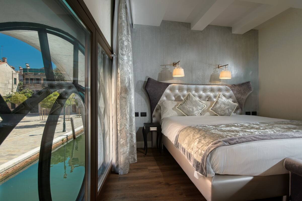 Ein Schlafzimmer mit großem Fenster, einem Bett mit geblattetem Kopfteil, zwei Nachttischlampen und Blick auf ein Gebäude und einen Pool.