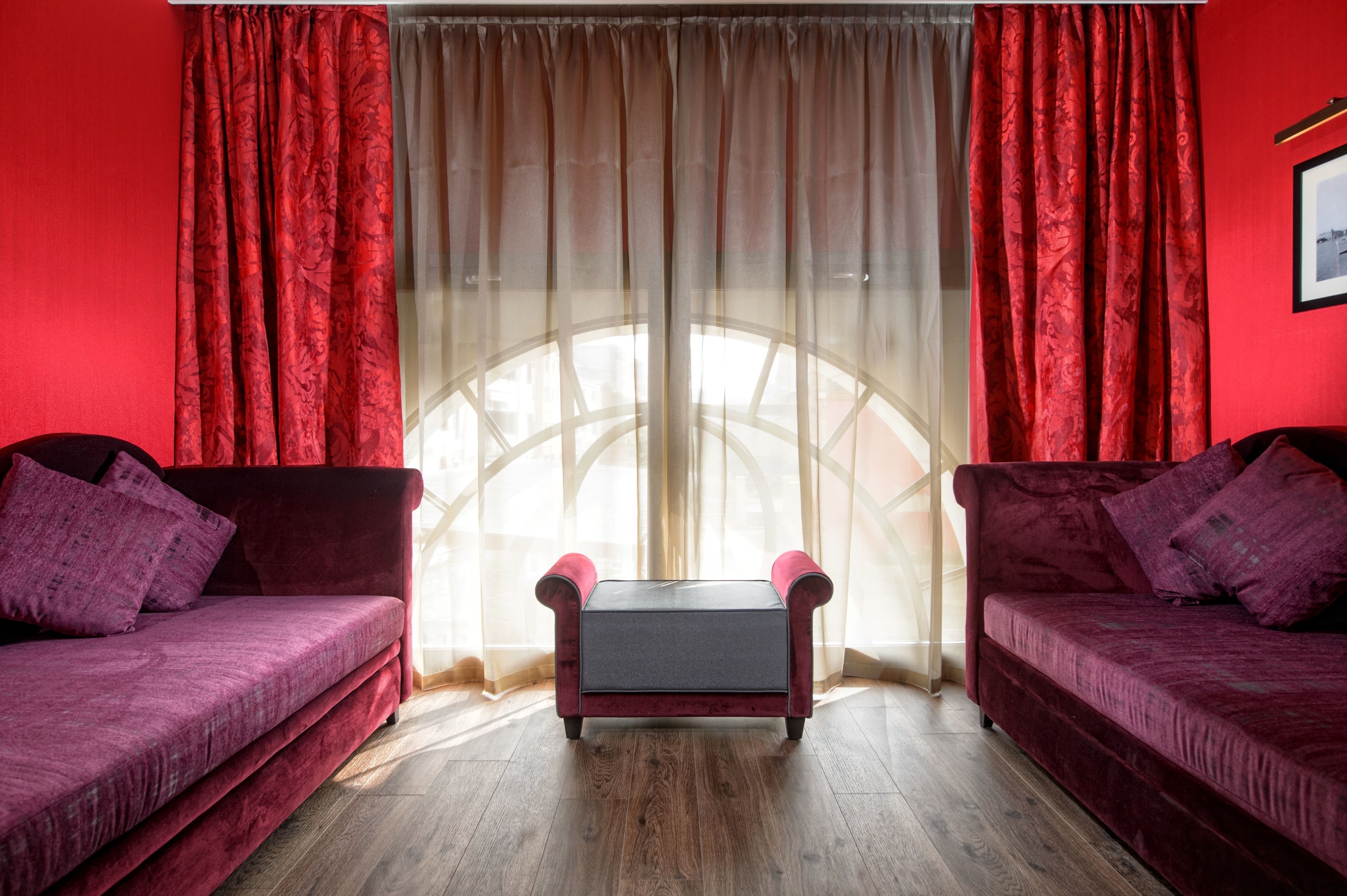 Quarto com dois sofás de veludo vermelho, uma mesa pequena e uma janela grande com cortinas leves.