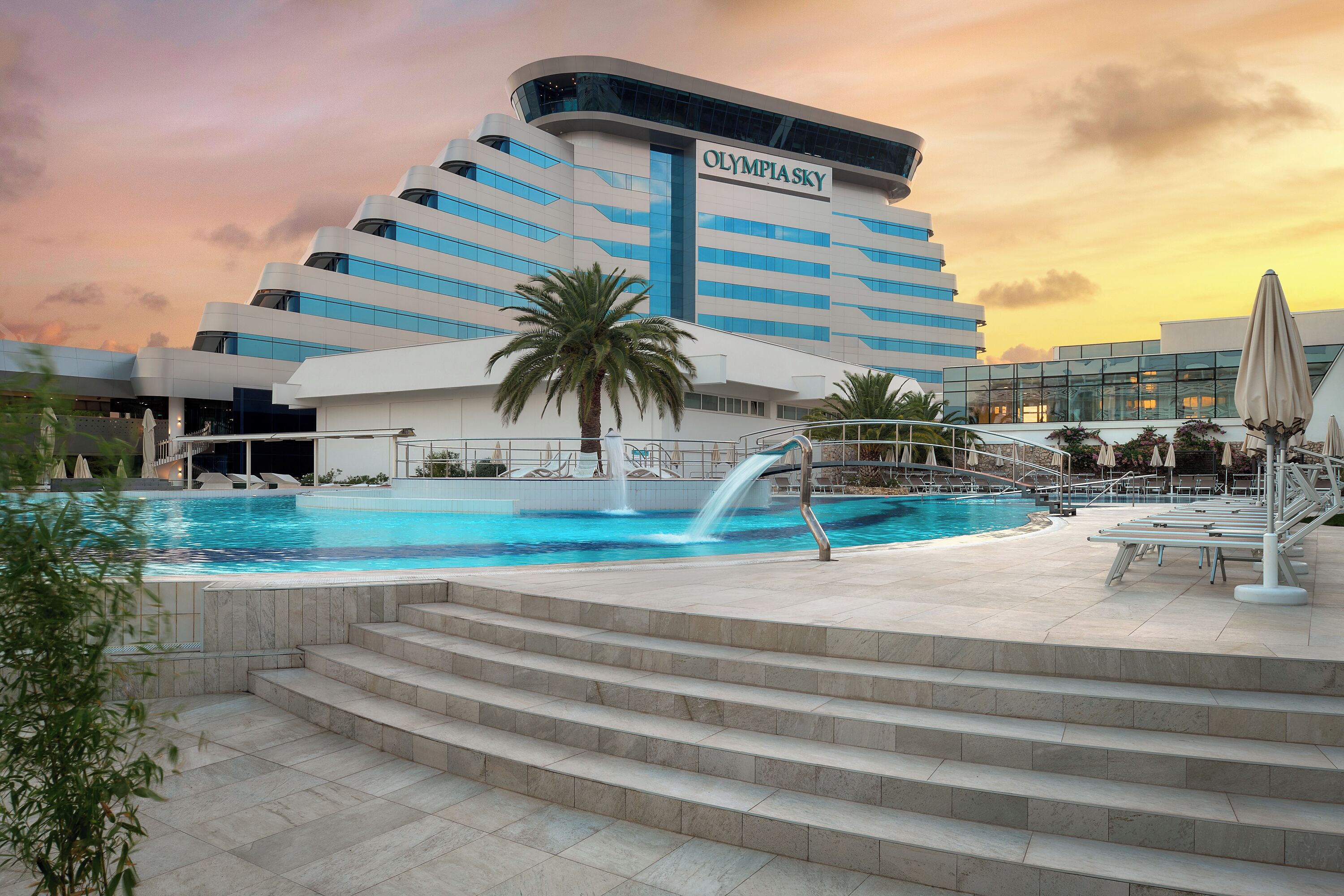 A modern hotel with a swimming pool, palm trees, and a sunset sky.