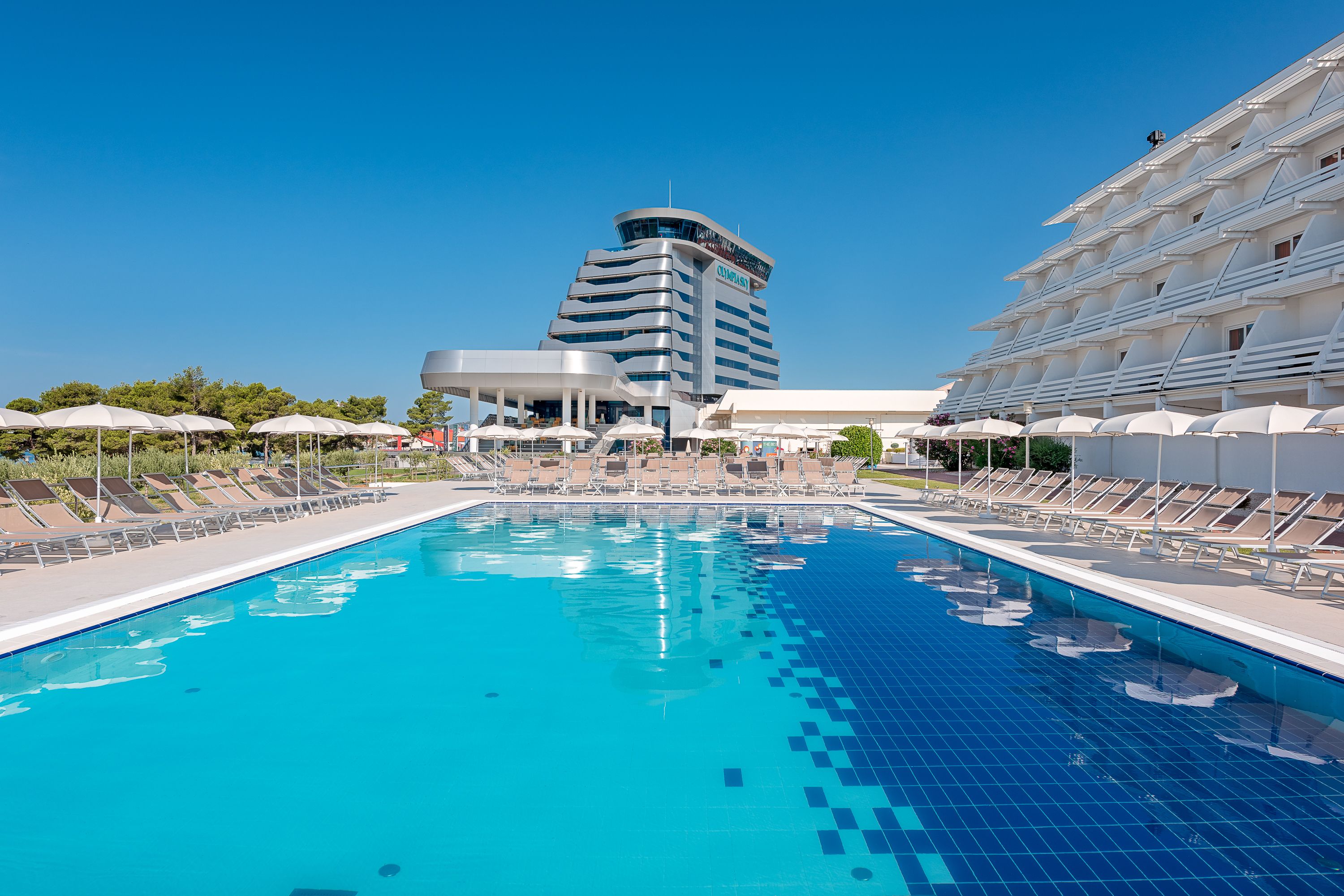 A swimming pool with lounge chairs and umbrellas, a modern building with a curved facade, and a clear blue sky.