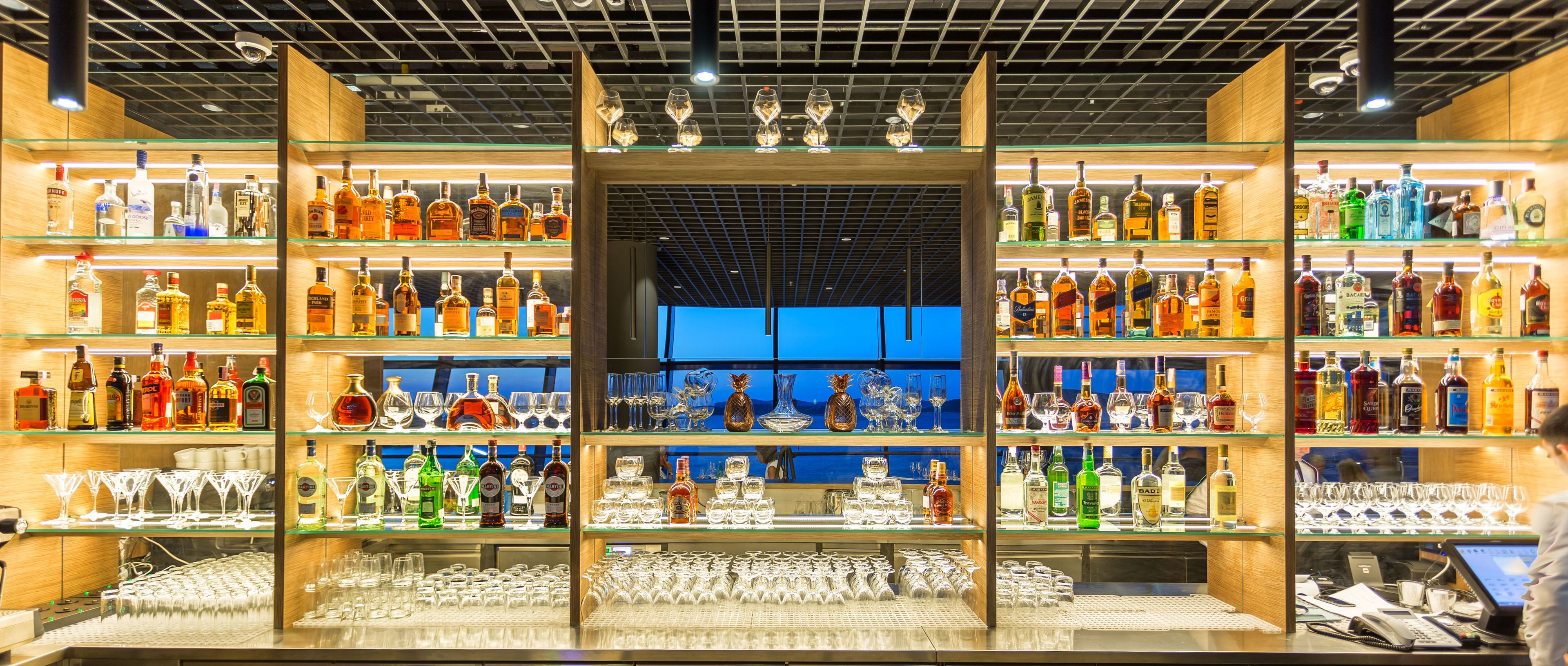 A bar with multiple shelves of liquor bottles and glasses, a laptop on the counter, and a view of the sky through the ceiling.
