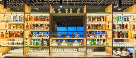 A bar with multiple shelves of liquor bottles and glasses, a laptop on the counter, and a view of the sky through the ceiling.