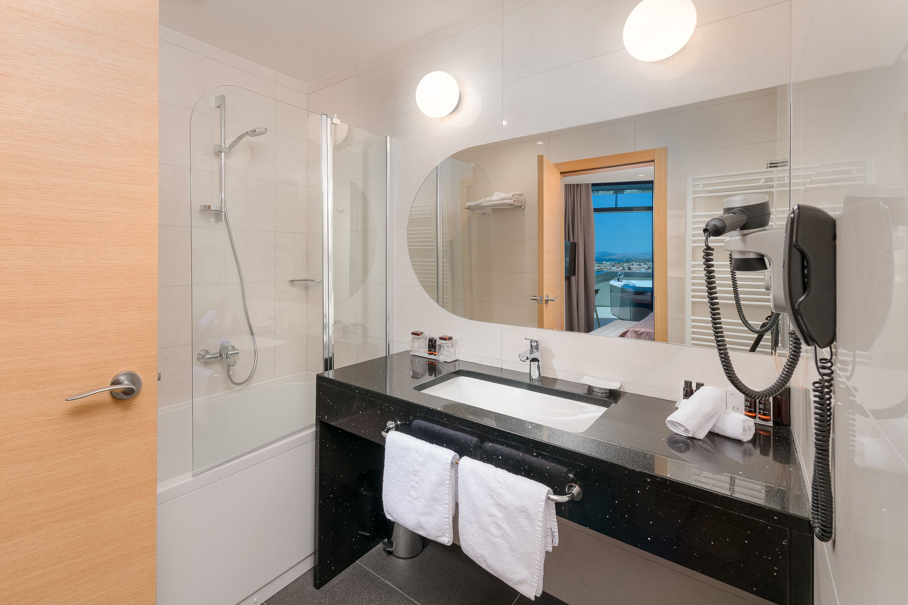 Un baño moderno con ducha envuelta en vidrio, lavamanos con encimera negra, un espejo grande y un secador de pelo en la pared.