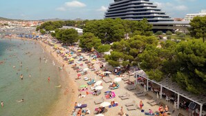 Uma praia com vários espreguiçadeiras e guarda-sóis, um edifício grande ao fundo e céu limpo.