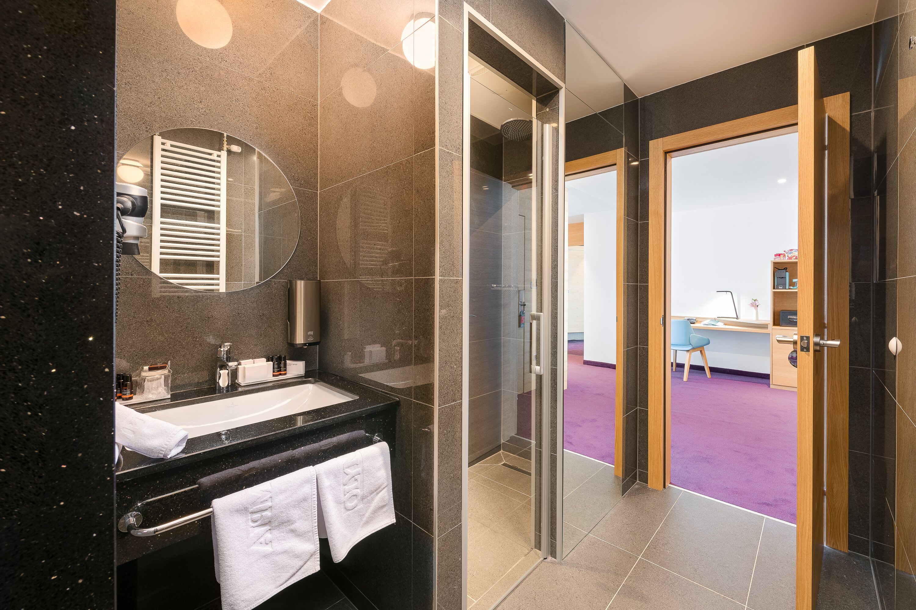 Ein modernes Badezimmer mit Duschkabine aus Glas, einem Waschbecken mit Spiegel und einem violetten Teppich, der durch die Tür sichtbar ist.