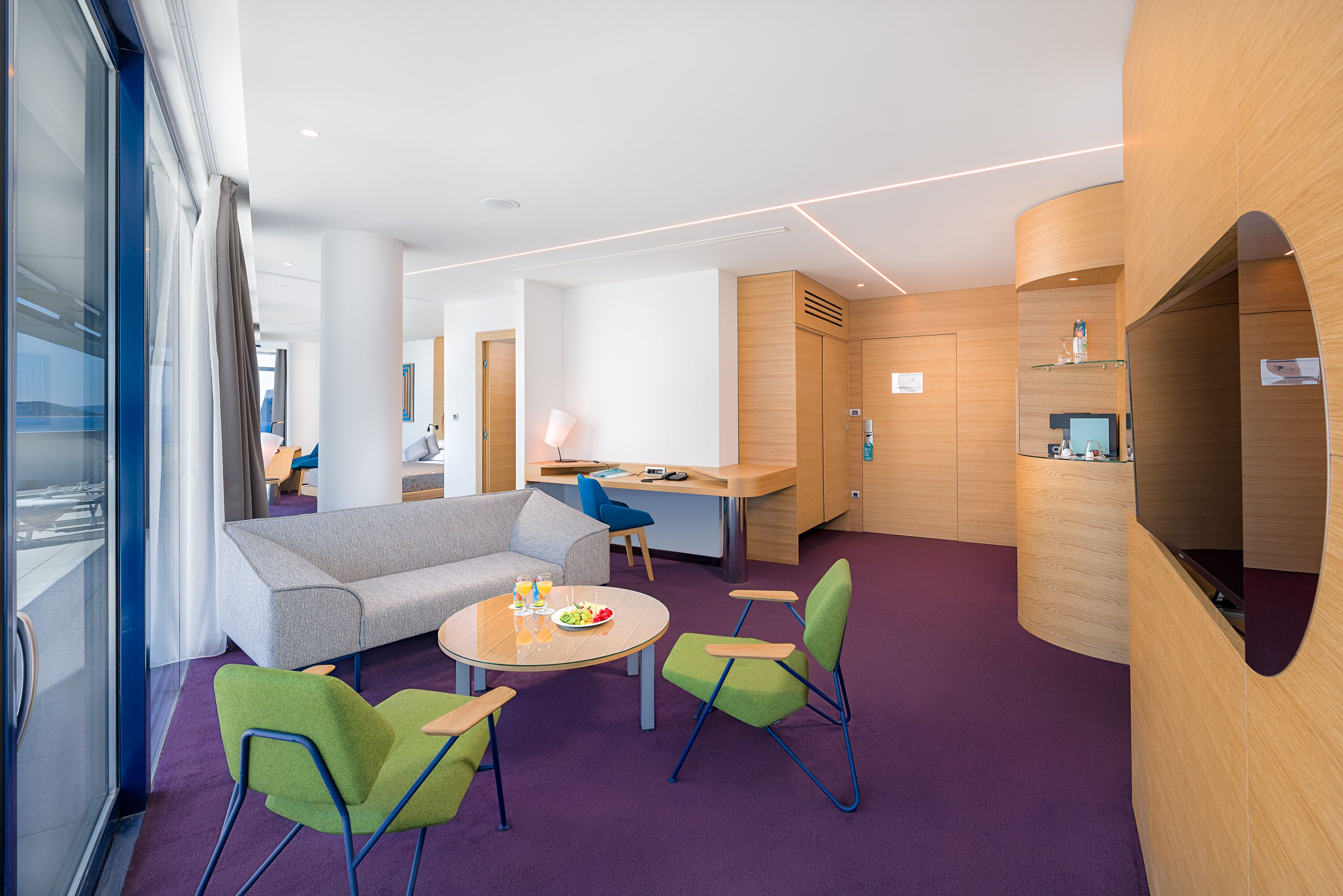 A modern hotel lobby with a purple carpet, a grey sofa, green chairs, a round table with a fruit bowl, and wooden accents.