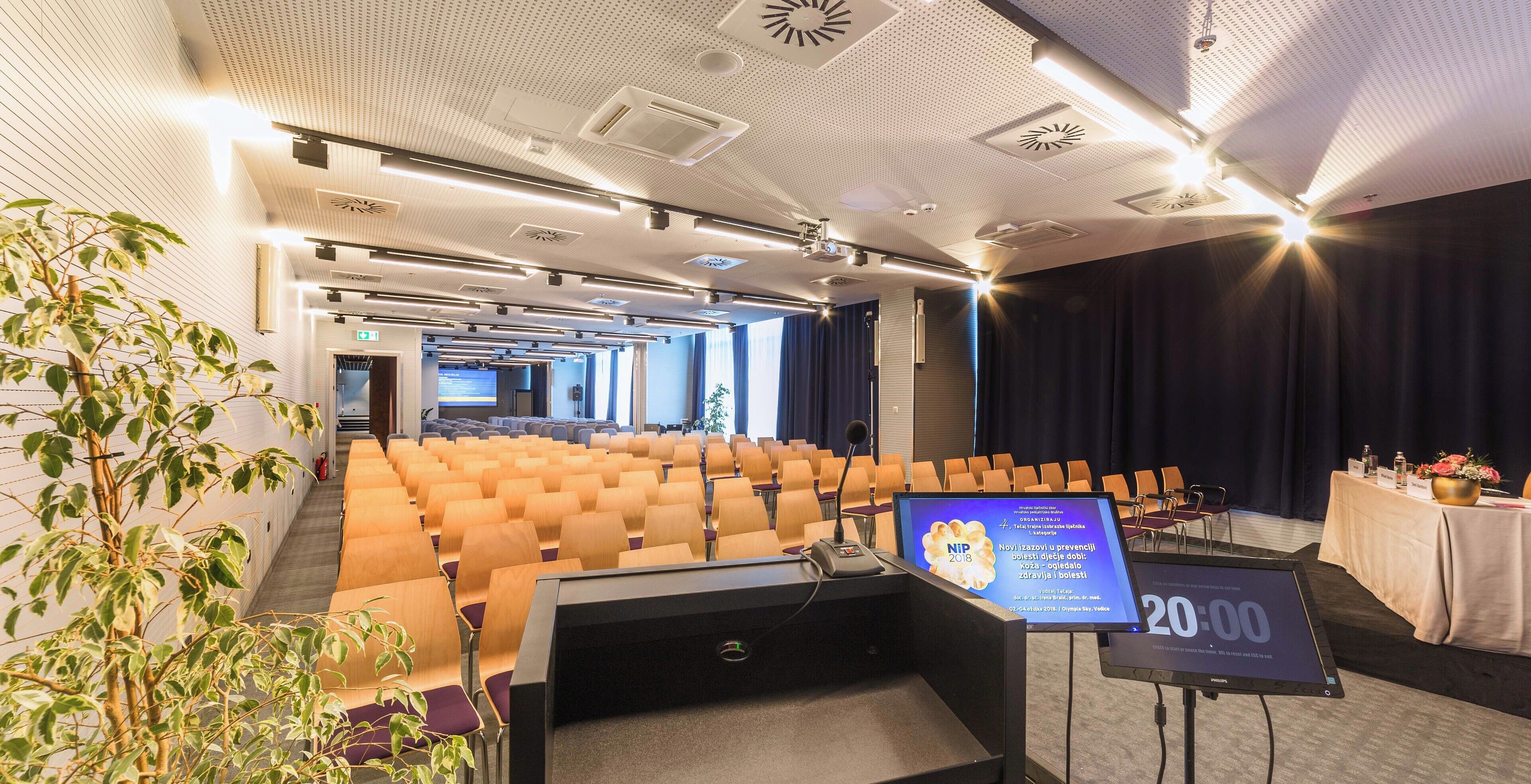 A conference room with rows of chairs, a podium, a large screen, and a clock displaying 20:00.