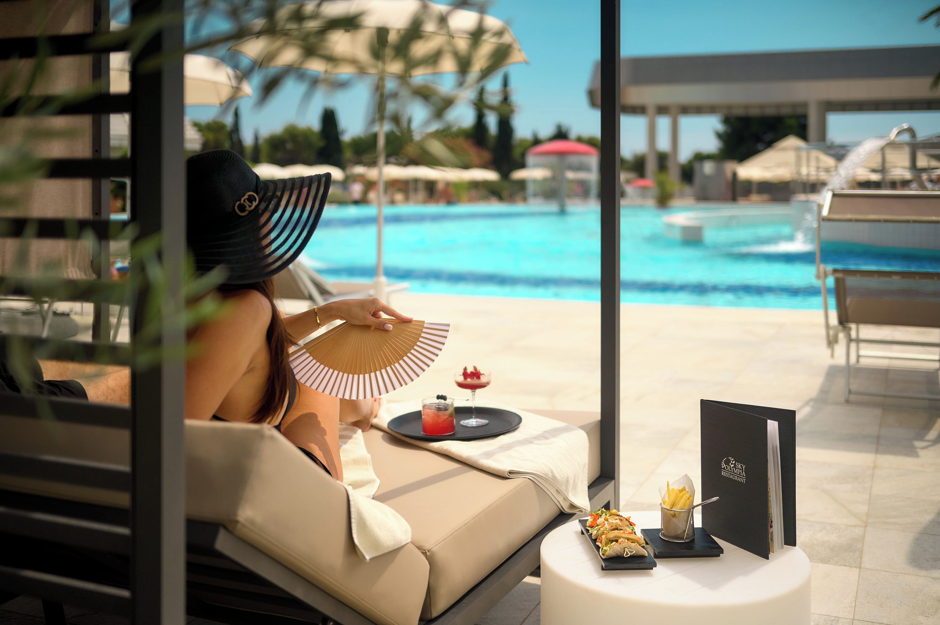 A person relaxing by a poolside lounge chair with a fan, a drink, and a menu on a small table.