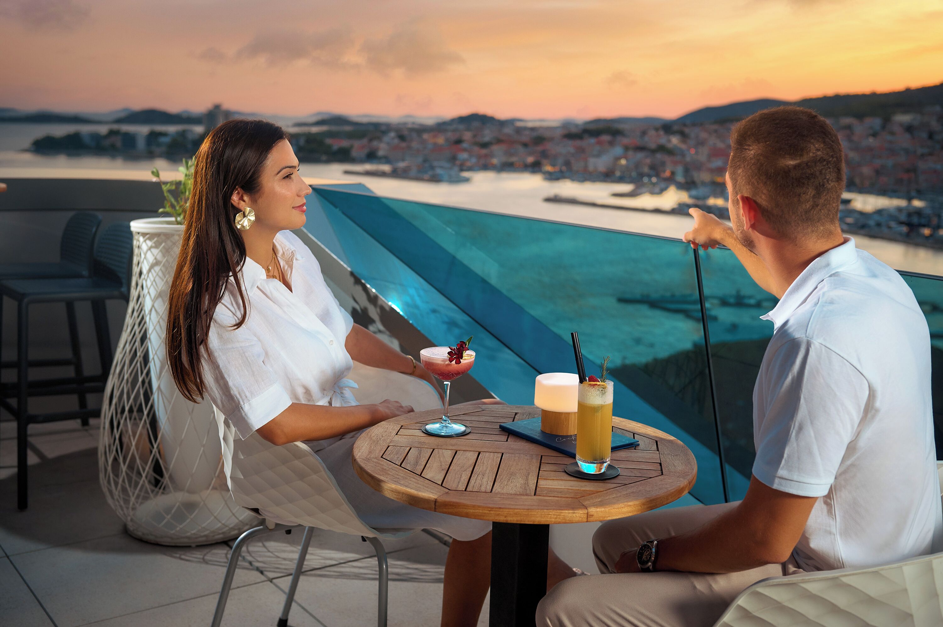Una pareja sentada en una mesa redonda de madera, con bebidas, con vista a una ciudad costera.