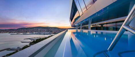 A modern building with a long, narrow pool extending from the main structure, overlooking a body of water and a distant cityscape.
