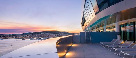 A modern building with a rooftop terrace featuring lounge chairs and a view of a cityscape at sunset.