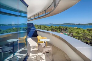 A balcony with a glass wall, yellow chairs, a small table, and a view of a body of water and greenery.