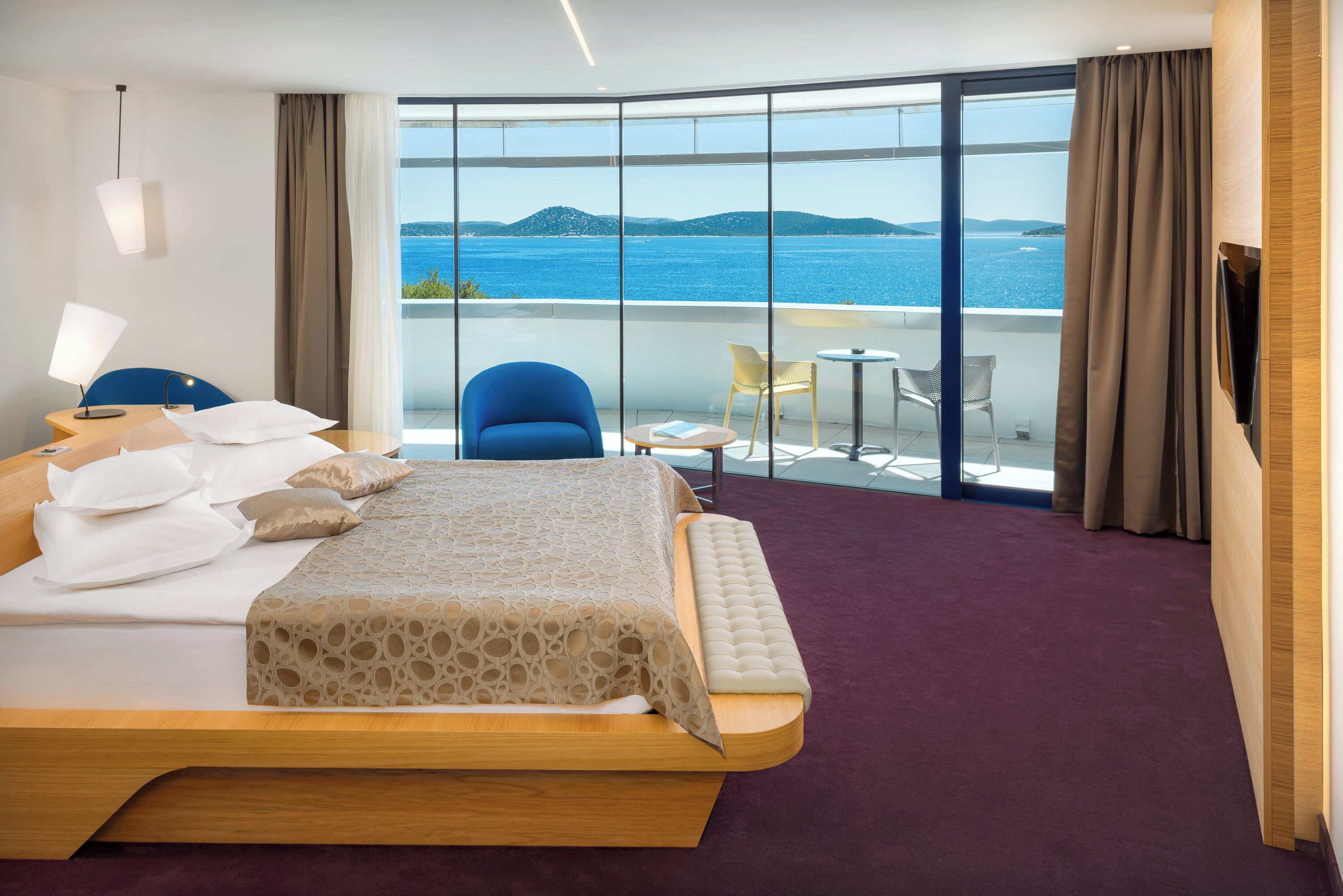 A hotel room with a large bed, a small balcony with a table and chairs, and a view of the sea.