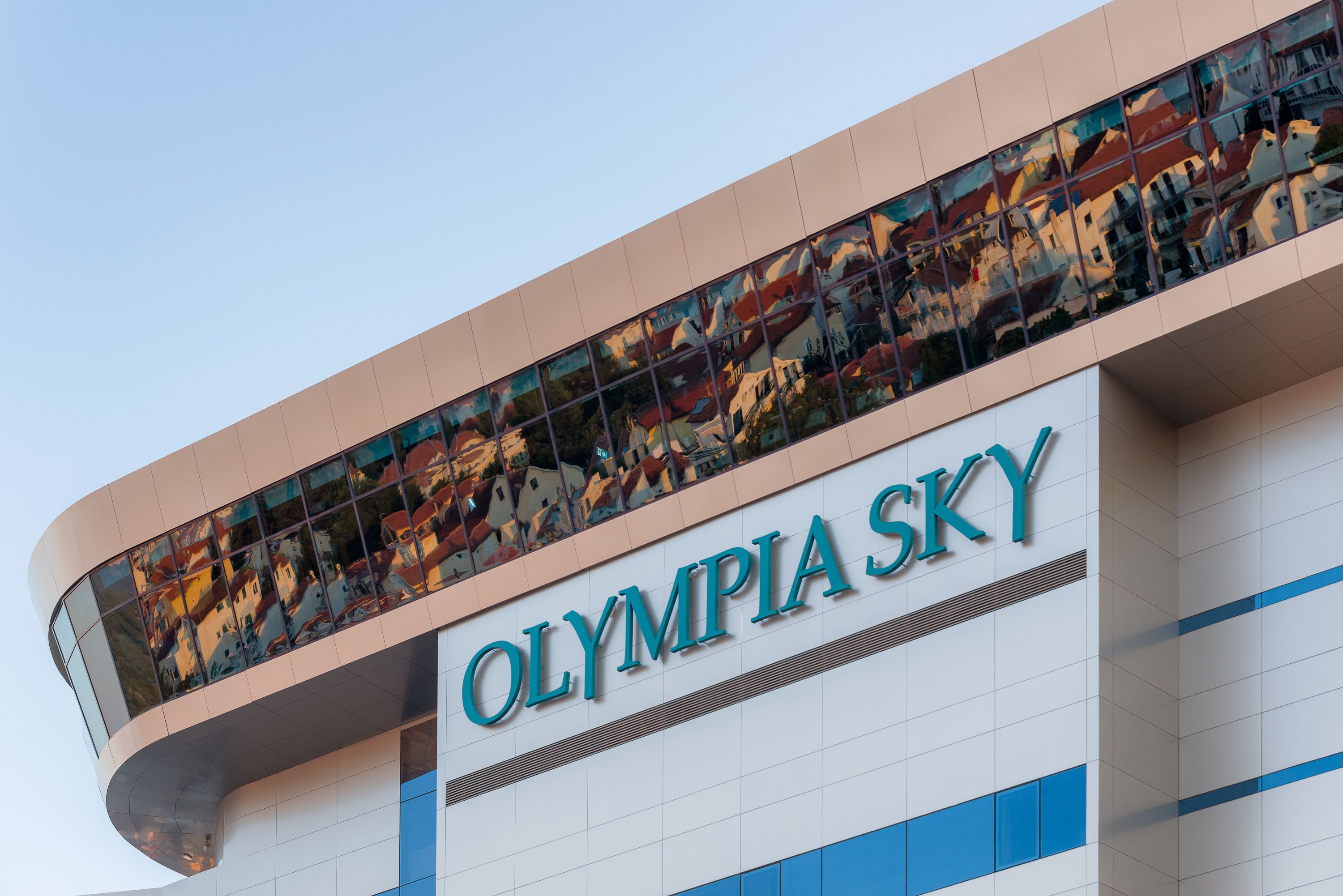 Rakennuksessa on kyltti, jossa lukee ”OLYMPIA SKY”, ja sen suuri lasijulkisivu heijastaa ympäröiviä rakennuksia.