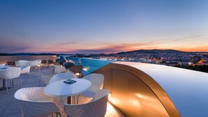 Terraço no telhado com cadeiras e mesas brancas, com vista para um horizonte urbano e uma praia ao pôr do sol.