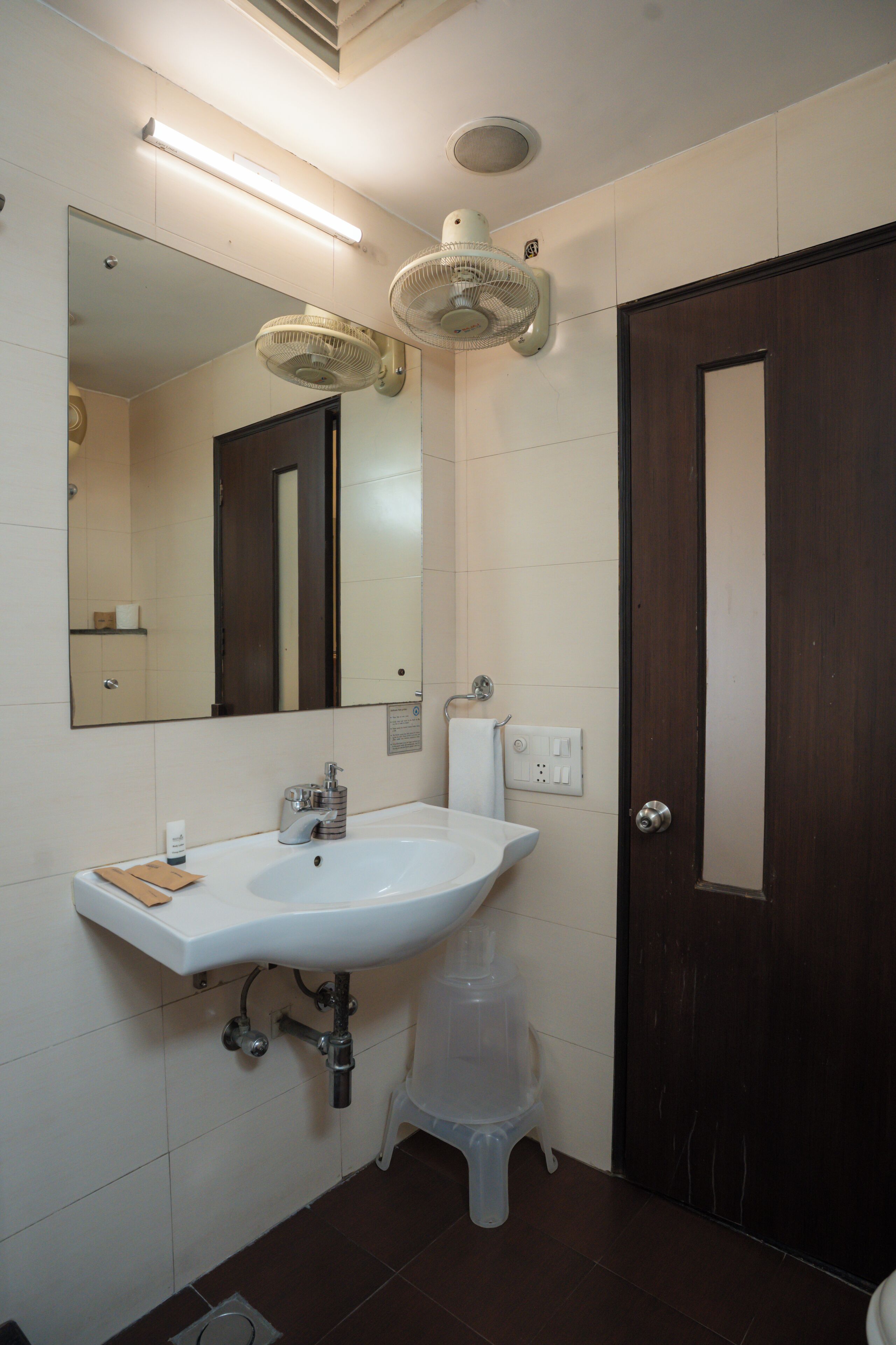 A bathroom with a white sink, a mirror, a wall-mounted fan, and a dark wooden door.