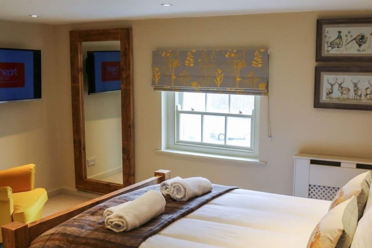 A neatly made bed with a rolled towel, a window with a floral blind, a mirror, and a framed picture on the wall.