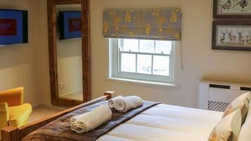 A neatly made bed with a rolled towel, a window with a floral blind, a mirror, and a framed picture on the wall.