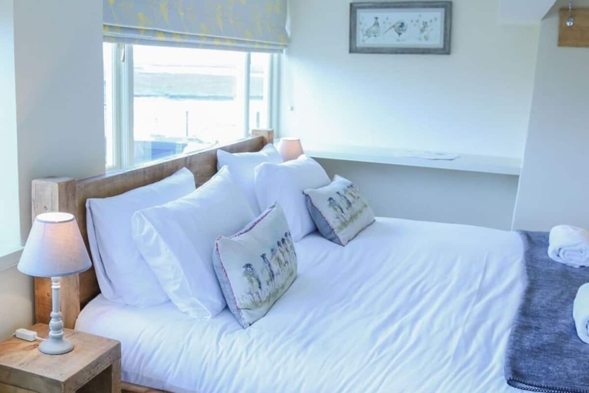 A neatly made bed with white linens and decorative pillows, a bedside lamp, and a window with a view of the outside.