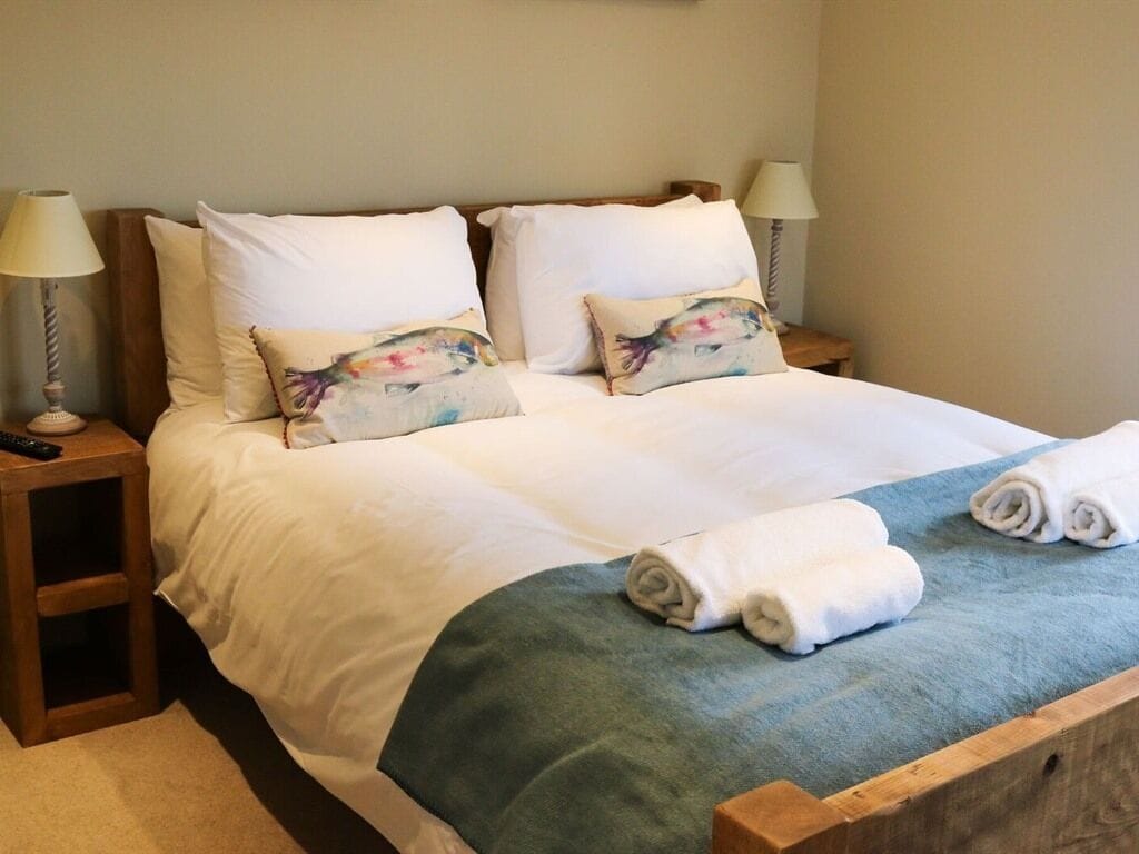 A neatly made bed with white linens, two decorative pillows, a blue throw blanket, and rolled towels on a wooden headboard.