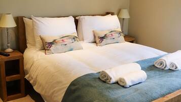 A neatly made bed with white linens, two decorative pillows, a blue throw blanket, and rolled towels on a wooden headboard.