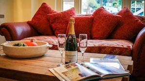 A cozy seating area with a wooden table, a bottle of champagne, two glasses, a bowl with a dish, and an open magazine. - The Quay B&B (Wells-next-the-Sea)