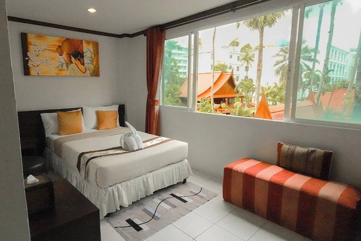 A hotel room with a bed, a bench, a painting, and a view of palm trees and buildings.