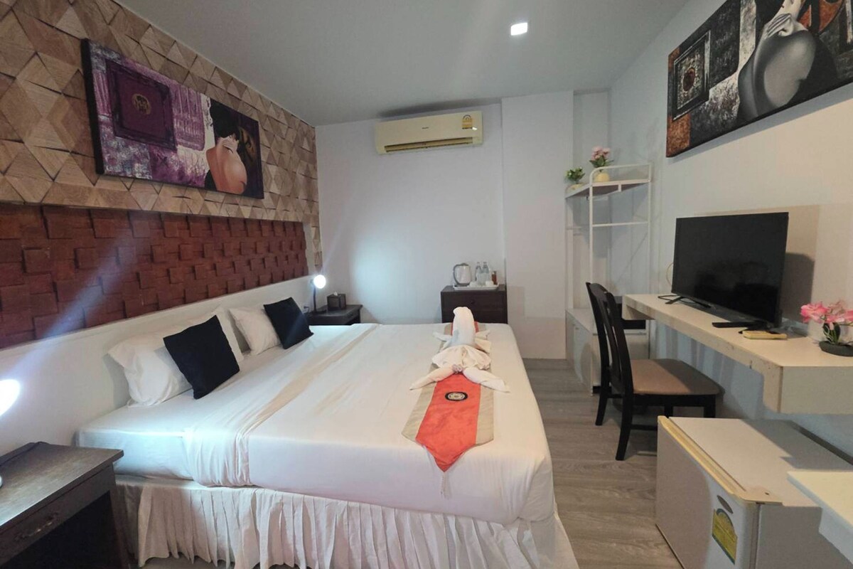 A hotel room with a bed, a desk with a TV, a chair, and a small table with a lamp.