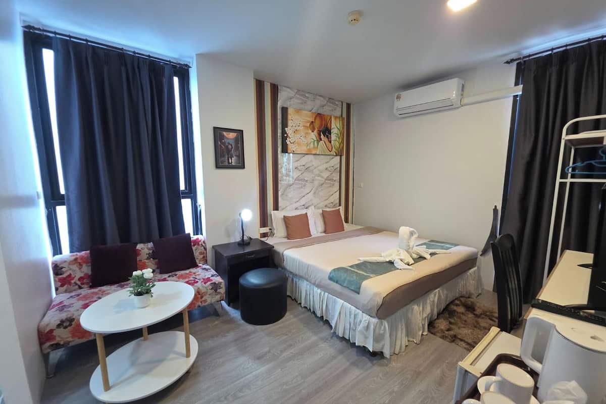 A hotel room with a bed, a sofa, a small table, a chair, a desk, and an air conditioning unit.
