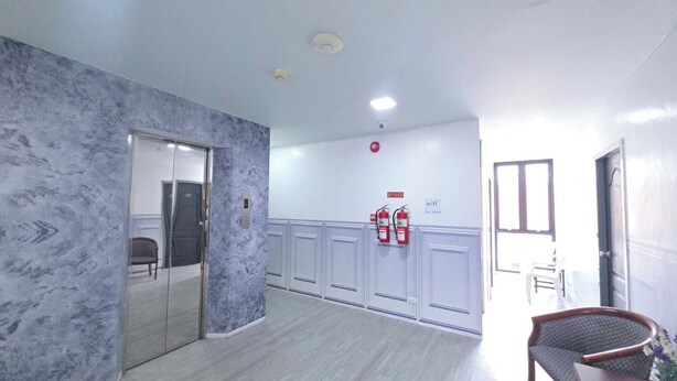 Un pasillo con pared de mármol, una puerta, una silla, una mesa con una planta y un extintor.