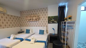 Habitación de hotel con dos camas, un escritorio con lámpara, un taburete pequeño, un espejo y una estantería con flores.
