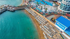An aerial view of a beach resort with a large pool area, multiple sun loungers, and a prominent building with a blue roof. - Granada Luxury Resort Okurcalar - All Inclusive (Alanya)