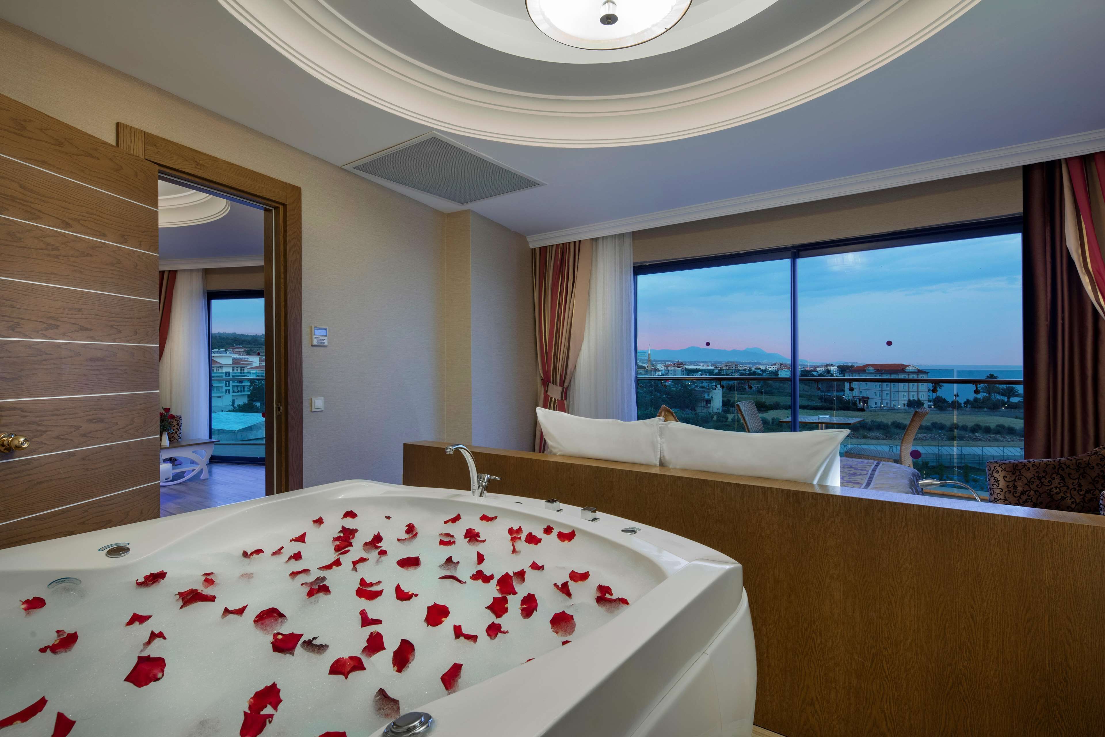 A modern hotel room with a large bathtub filled with rose petals, a view of the cityscape through large windows, and a cozy seating area with a view.