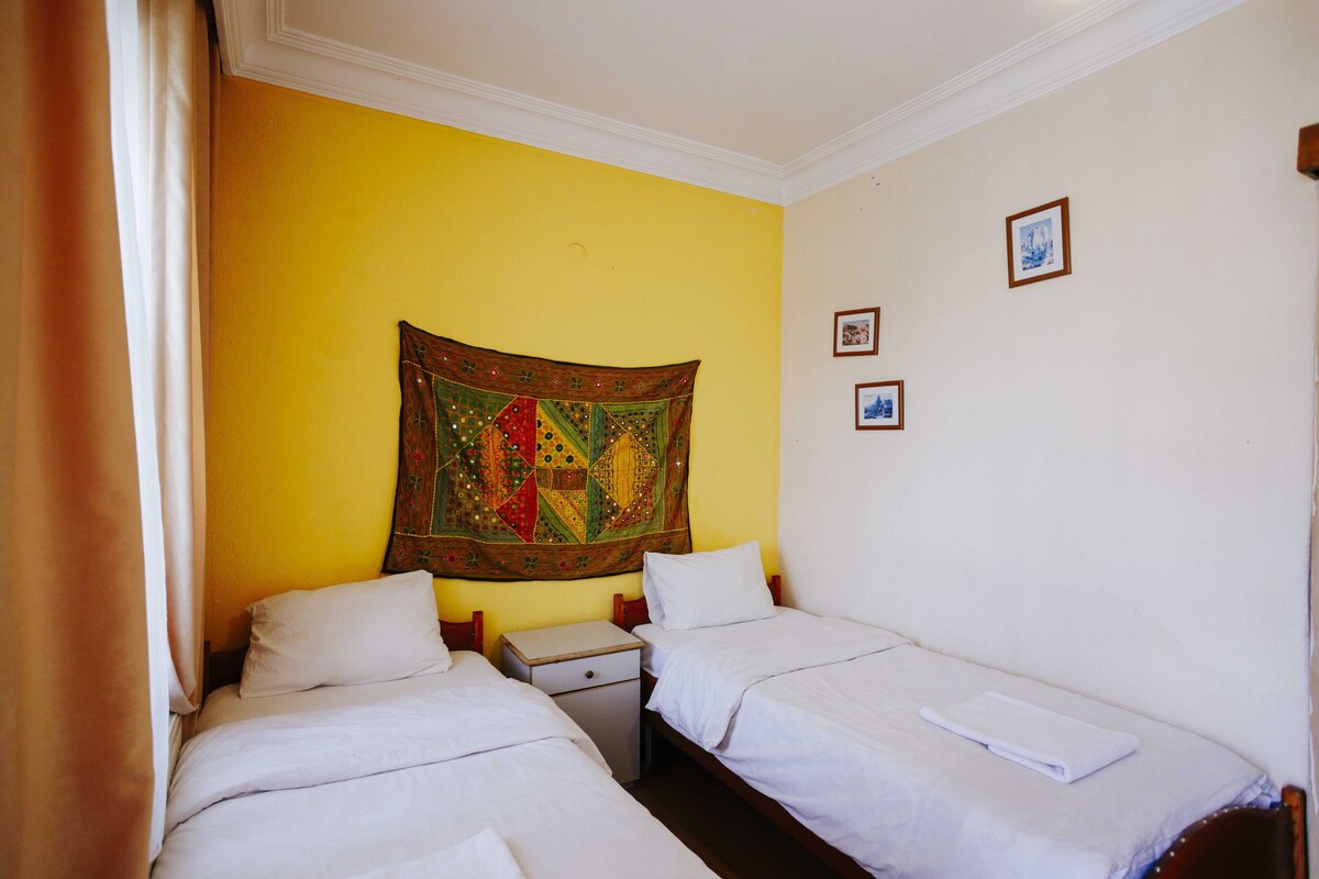 A hotel room with two single beds, a small bedside table, a tapestry on the wall, and a few framed pictures.
