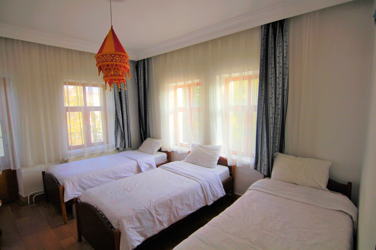A hotel room with two single beds, a hanging orange light fixture, and large windows with sheer curtains.