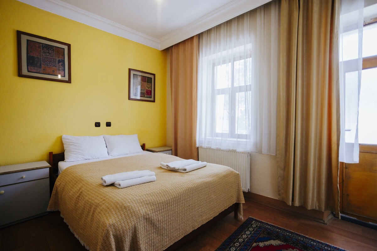 A hotel room with a bed, two framed pictures on the wall, a window with curtains, and a radiator.