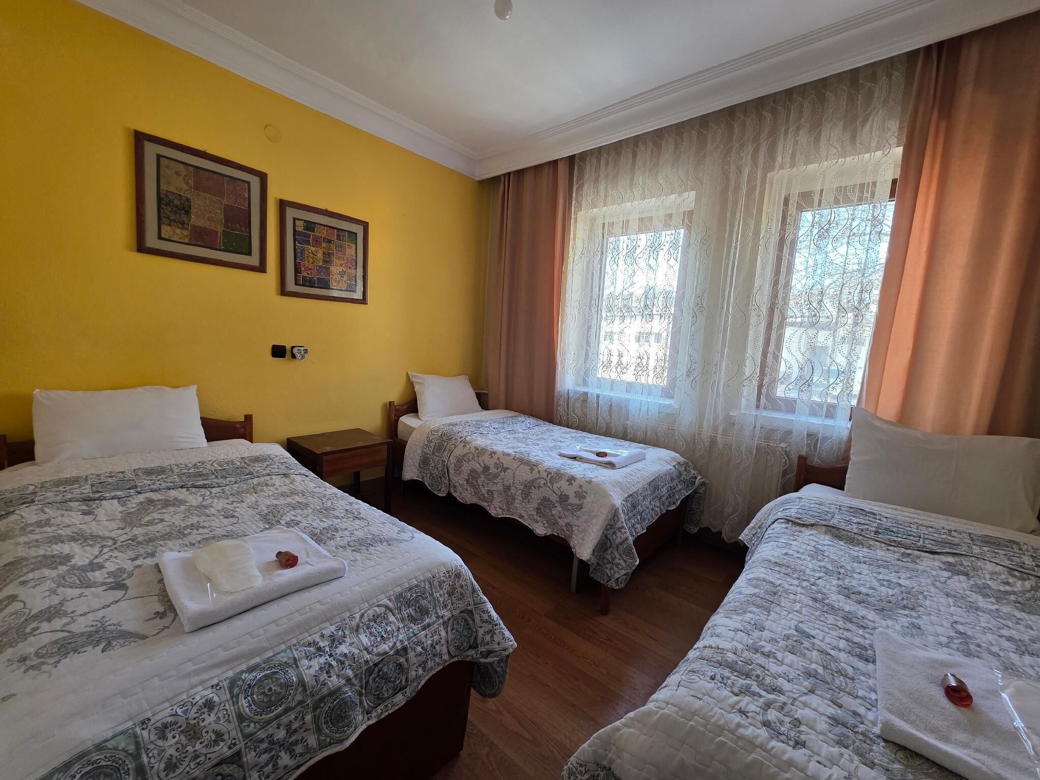 Habitación de hotel con tres camas, piso de madera, pared amarilla y dos cuadros enmarcados.