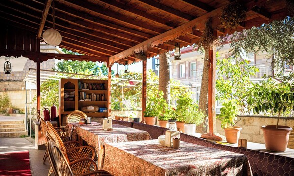 An outdoor dining area with wooden tables and chairs, potted plants, and a bookshelf. - Kose Pension (Nevsehir)