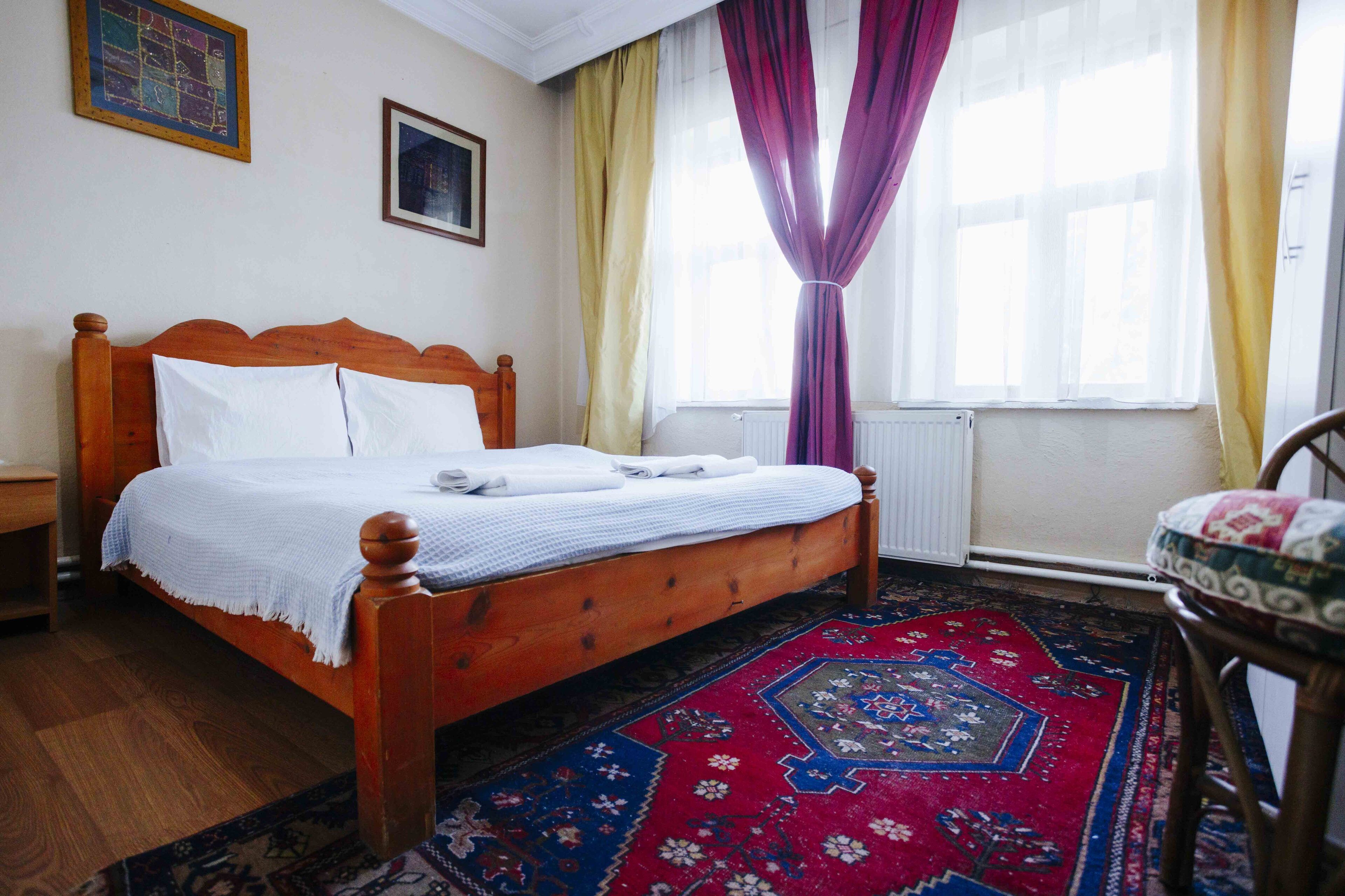 Uma cama bem arrumada com lençóis brancos, cabeceira de madeira e um tapete com padrão vermelho e azul no chão.