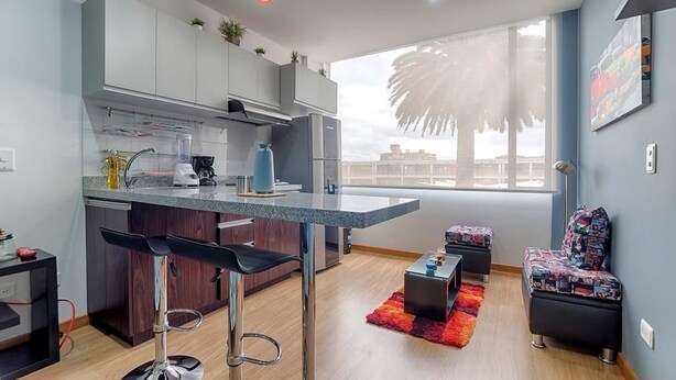 A modern kitchen with grey cabinets, a bar stool, a coffee machine, and a large window with a view of palm trees. - Las Palmas Apartment 306 (Bogotá)