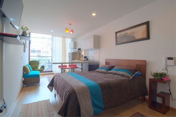 Ein modernes Hotelzimmer mit einem großen Bett, einer kleinen Küchenzeile, einer Sitzecke mit einem Sessel und einem Fenster mit Aussicht.