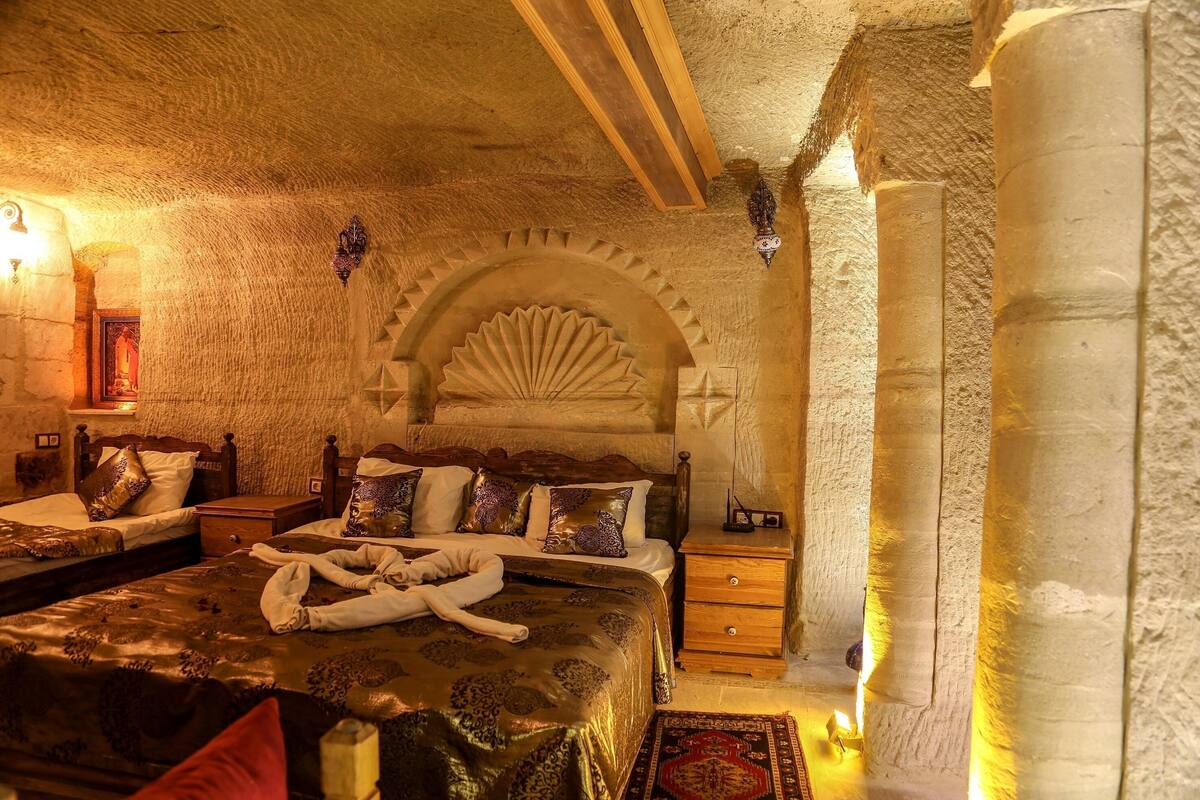 Um quarto com teto de pedra, cama com cabeceira decorativa, mesa de cabeceira de madeira e luminária fixada na parede.