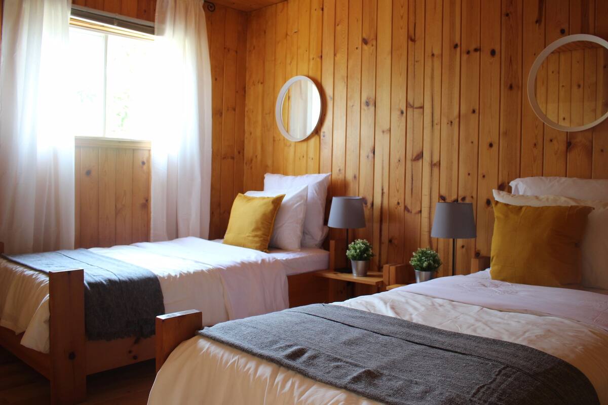 Ein Zimmer mit Holzboden und zwei Betten, jeweils mit weißer Bettwäsche und gelben Kissen. Zwei Nachttische mit Lampen und kleinen Topfpflanzen sind ebenfalls vorhanden.