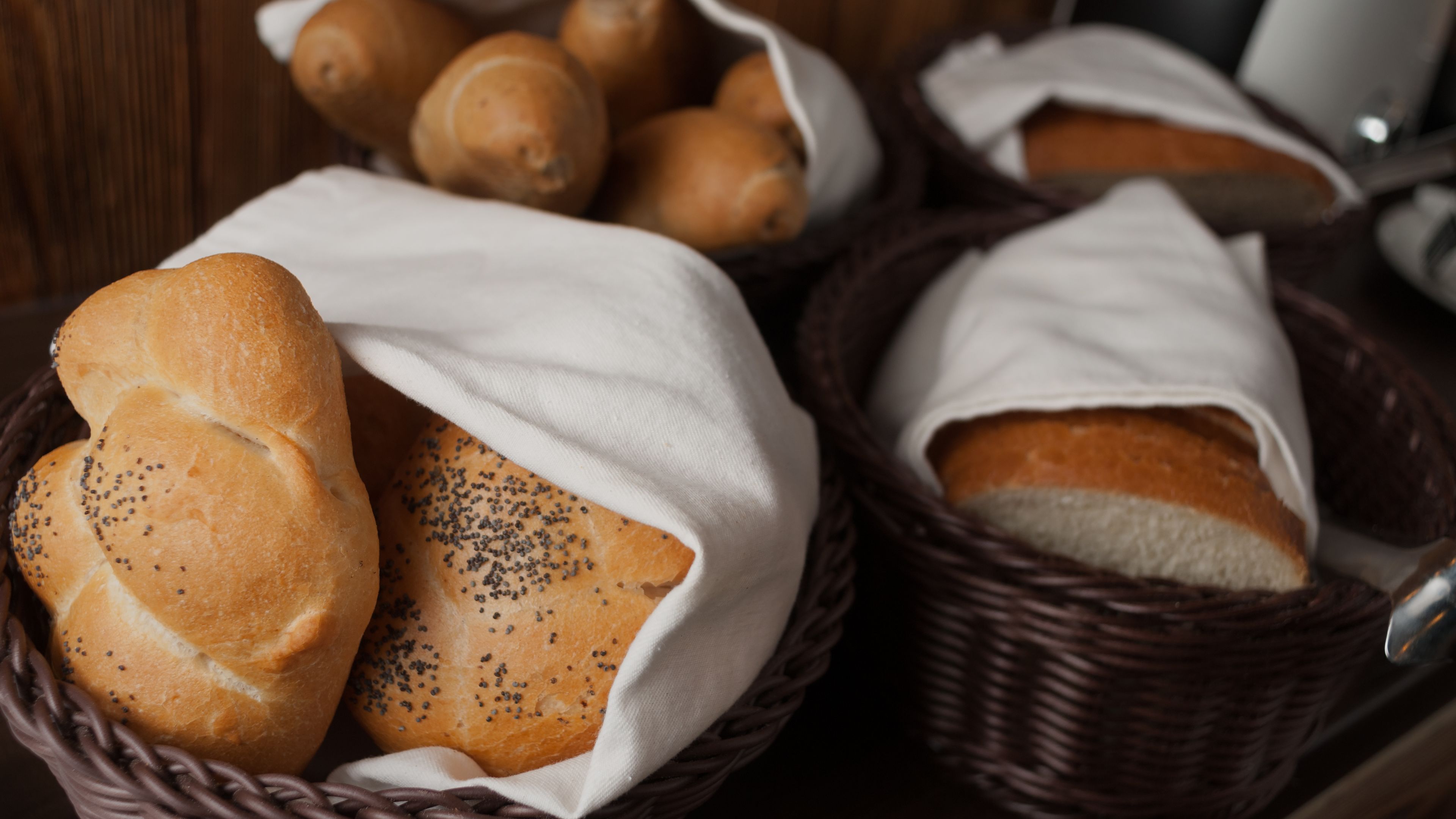 Ein Korb mit Brot mit Mohn und ein Korb mit Brötchen, beides in weißes Tuch gewickelt.