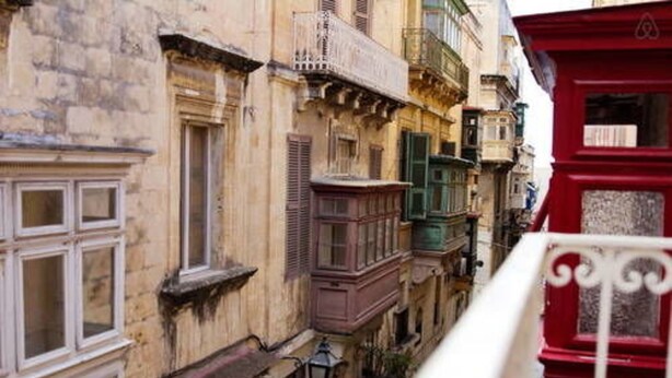 A narrow street with old buildings, one with a red facade and white balcony railing. - VBL Hostel – Adults only (Valletta)