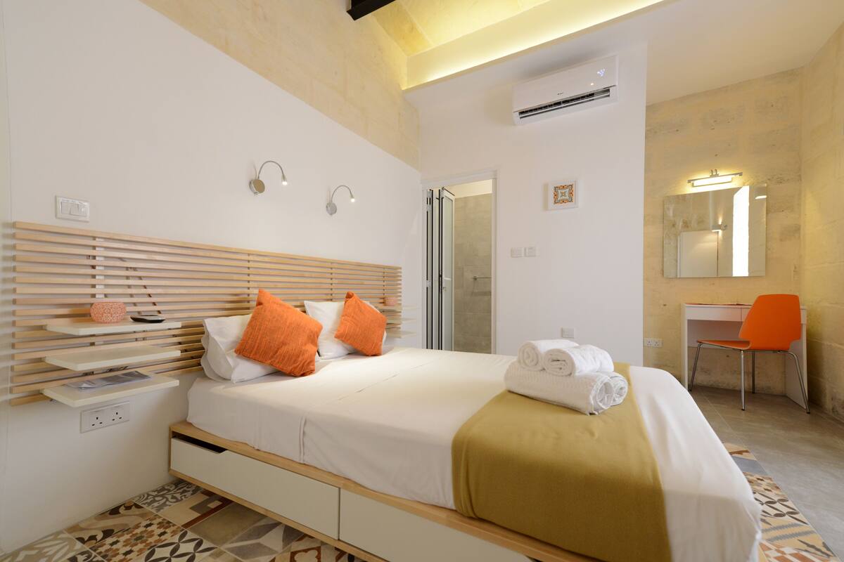 Ein modernes Hotelzimmer mit einem Holz-Kopfteil, einem Bett mit weißer Bettwäsche und orangefarbenen Kissen, einem Schreibtisch mit Stuhl sowie einem Badezimmer mit Spiegel und Dusche.
