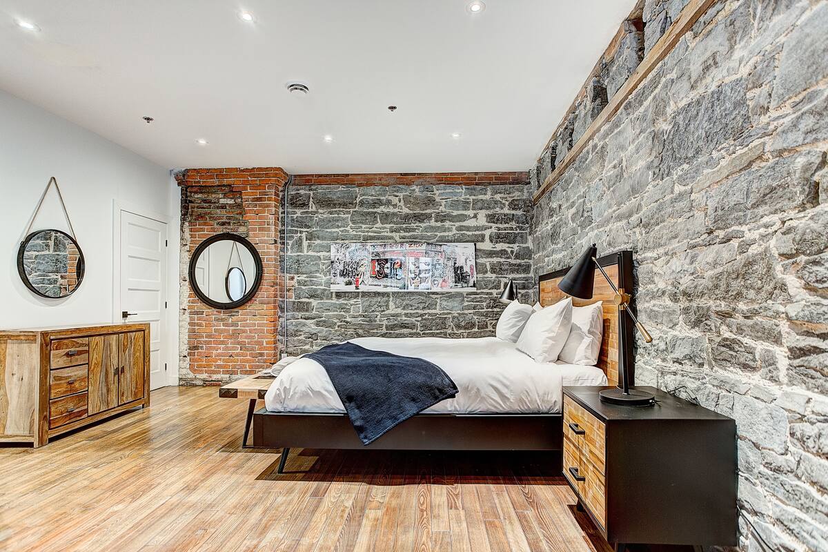 Un dormitorio con pared de ladrillo, cómoda de madera, cama con cabecera negra y mesita de noche con lámpara.