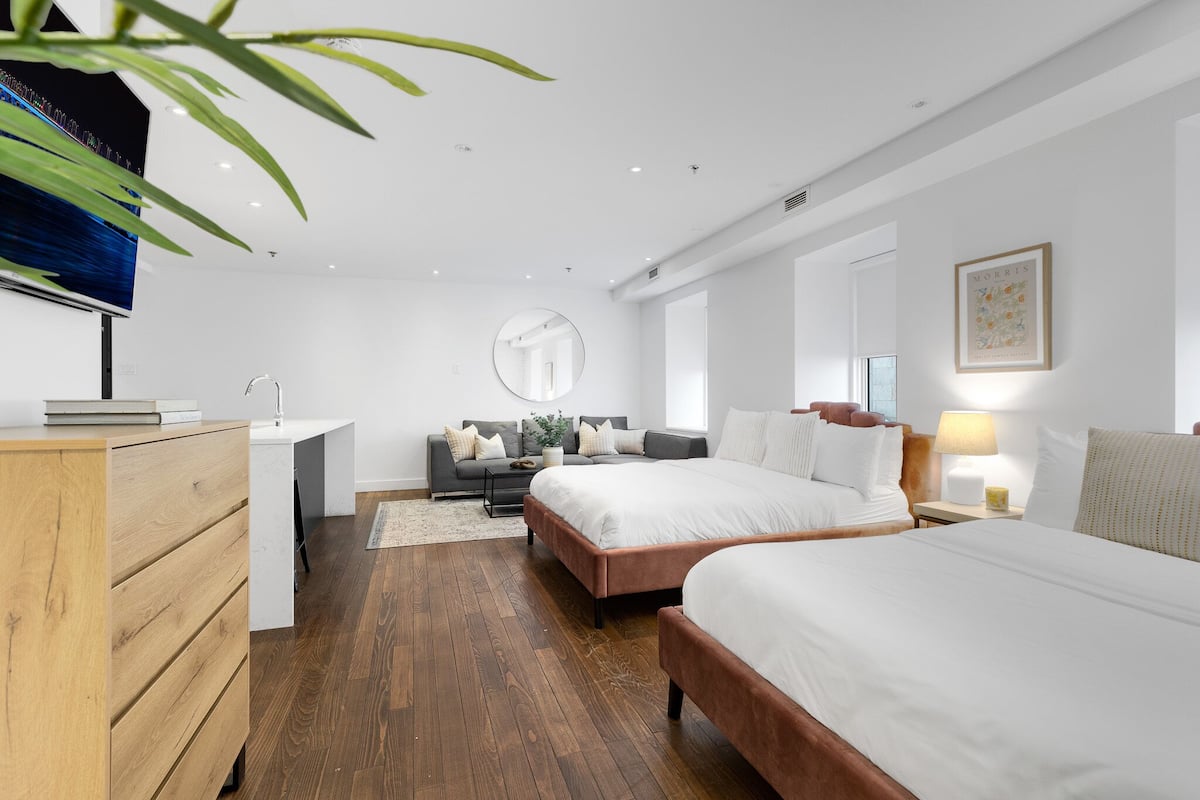 Una habitación de hotel moderna con dos camas, un aparador de madera, un espejo redondo, un sofá y un televisor.