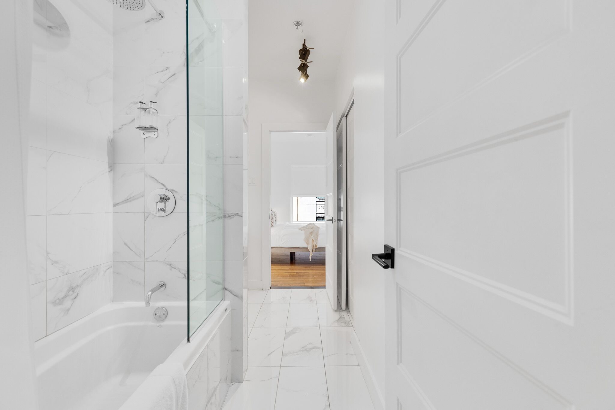 A modern bathroom with a glass shower enclosure, a white bathtub, and a hallway leading to a bedroom.