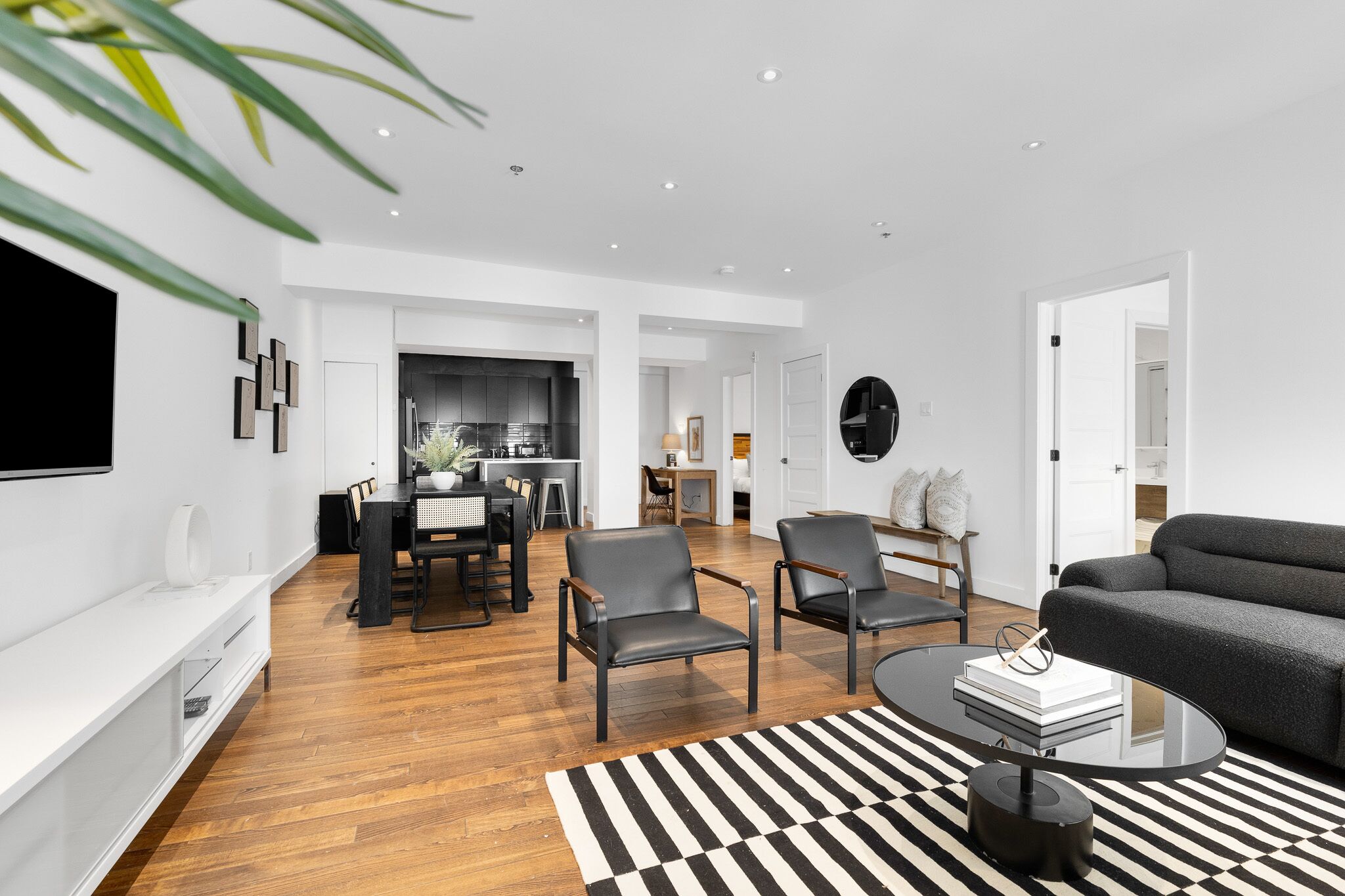 A modern living room with a black and white striped rug, a black coffee table, and a grey sofa. There is a dining area with a table and chairs, and a kitchen in the background.